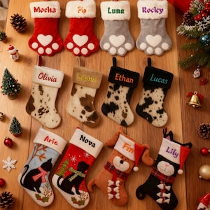 Op de afbeelding: Een verzameling gepersonaliseerde kerstsokken in verschillende ontwerpen. De sokken hebben namen als "Mocha", "Pip" en "Luna". Sommige hebben pootafdrukontwerpen, andere dierenprints of stripfiguren. Elke sok heeft een witte manchet.