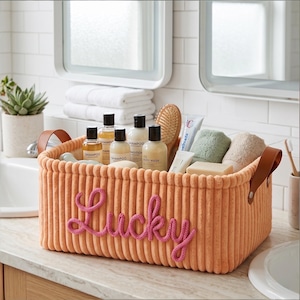 Könnte beinhalten: Ein orangefarbener Korb aus Cord mit dem Wort "Lucky" aus rosa Garn. Der Korb enthält Toilettenartikel, darunter Shampoo, Spülung, Duschgel, eine Bürste und Handtücher. Der Korb hat braune Ledergriffe.