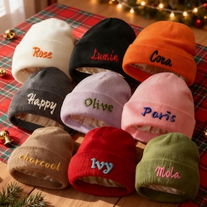 Puede incluir: Una colección de gorros de punto de varios colores: blanco, negro, naranja, gris, lavanda, rosa, marrón, rojo y verde. Cada gorro tiene un nombre bordado en un color contrastante. Los gorros están forrados con un material similar al satén.