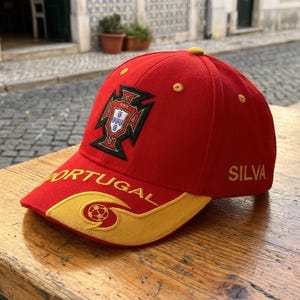 May include: A red baseball cap with a yellow brim and accents. The cap features the word "PORTUGAL" in white, a crest, and the name "SILVA". The cap is sitting on a wooden table.