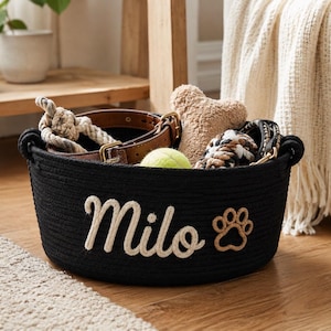 Peut inclure: Panier en corde noir avec le nom "Milo" en &eacute;criture cr&egrave;me et une empreinte de patte. Le panier contient un collier pour chien, un jouet en corde, une balle de tennis et d'autres accessoires pour chien. Le panier est sur un sol en bois.