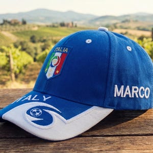 May include: A blue baseball cap with the word "MARCO" embroidered in white. The cap features the Italian flag and the word "ITALY". The brim is white with a soccer ball design. The cap is sitting on a wooden surface.