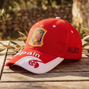 May include: A red baseball cap with a white brim, featuring the word "Spain" and a soccer ball graphic. The cap has the Spanish coat of arms and the name "ALEX" on the side. The cap is on a wooden table.