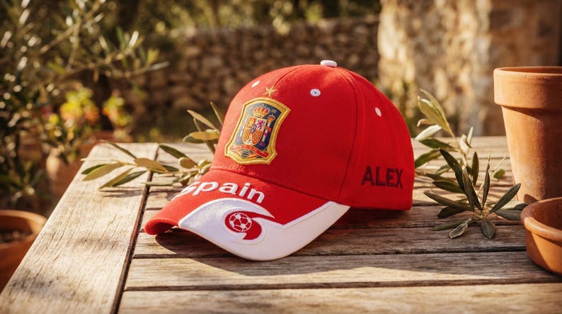 May include: A red baseball cap with a white brim, featuring the word "Spain" and a soccer ball graphic. The cap has the Spanish coat of arms and the name "ALEX" on the side. The cap is on a wooden table.