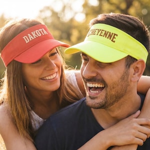 May include: A couple wearing visors with the names "DAKOTA" and "CHEYENNE" printed on them. The visor on the left is red, and the visor on the right is yellow. The couple is smiling and embracing outdoors.