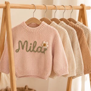 Könnte beinhalten: Eine Reihe von fünf langärmeligen Strickpullovern, die an einem Holzständer hängen. Der erste Pullover ist hellrosa mit dem Namen Mila in grüner Schreibschrift und einer weißen Gänseblümchen-Applikation. Die anderen vier sind in Creme- und Beigetönen.