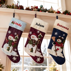May include: Three Christmas stockings hang from a mantel. Each stocking is a different color: burgundy, navy blue, and light blue. Each stocking has a snowman design and a name embroidered on the cuff: Linda, Adam, and Susanna.
