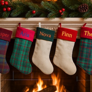 Puede incluir: Cinco calcetines navideños cuelgan sobre una chimenea. Cada calcetín tiene un nombre bordado en el puño. Los calcetines son de varios colores y estampados, incluyendo cuadros escoceses y blanco liso. Una guirnalda de vegetación y bayas decora la repisa de la chimenea.