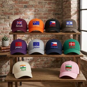 Könnte beinhalten: Eine Auswahl an Baseballkappen in verschiedenen Farben, jede mit einer Flagge und einem Namen bestickt. Die Kappen sind auf Holzregalen vor einer Backsteinmauer angeordnet. Die Flaggen repräsentieren verschiedene Länder, und die Namen sind in Kursivschrift.