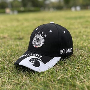 May include: Black baseball cap with the German national team emblem, white stars, and the name "SCHMIDT". The bill is white with a soccer ball design and the word "Germany".