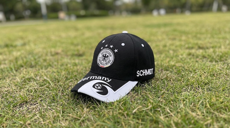 May include: Black baseball cap with the German national team emblem, white stars, and the name "SCHMIDT". The bill is white with a soccer ball design and the word "Germany".