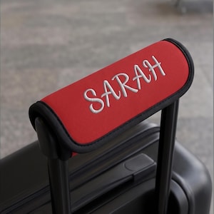 May include: A bright red luggage handle wrap featuring the name SARAH embroidered in white thread. The rectangular wrap has black trim along the edges and is securely fastened around the black telescopic handle of a piece of rolling luggage.