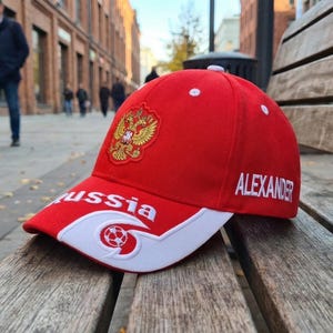 May include: Red baseball cap with white accents, featuring the Russian coat of arms and the name "ALEXANDER" embroidered in white. The word "Russia" is also displayed. The cap is resting on a wooden bench.