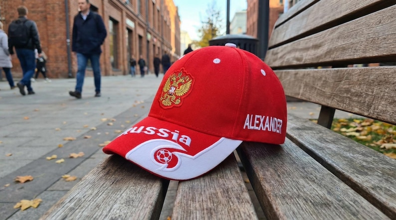 May include: Red baseball cap with white accents, featuring the Russian coat of arms and the name "ALEXANDER" embroidered in white. The word "Russia" is also displayed. The cap is resting on a wooden bench.