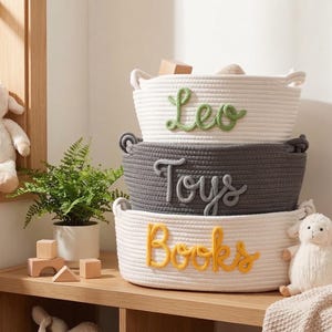 Peut inclure: Trois paniers de rangement tress&eacute;s empil&eacute;s sur une &eacute;tag&egrave;re en bois. Le panier sup&eacute;rieur est blanc avec "Leo" en &eacute;criture verte. Le panier du milieu est gris avec "Toys" en &eacute;criture grise. Le panier inf&eacute;rieur est blanc avec "Books" en &eacute;criture jaune.