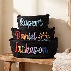 Peut inclure: Trois paniers noirs en corde empil&eacute;s, chacun avec un nom en lettres color&eacute;es : Rupert, Daniel et Jackson. Le panier Daniel a un motif de soleil et de vagues. Une paire de petites chaussures et une couverture pli&eacute;e sont sur la table.