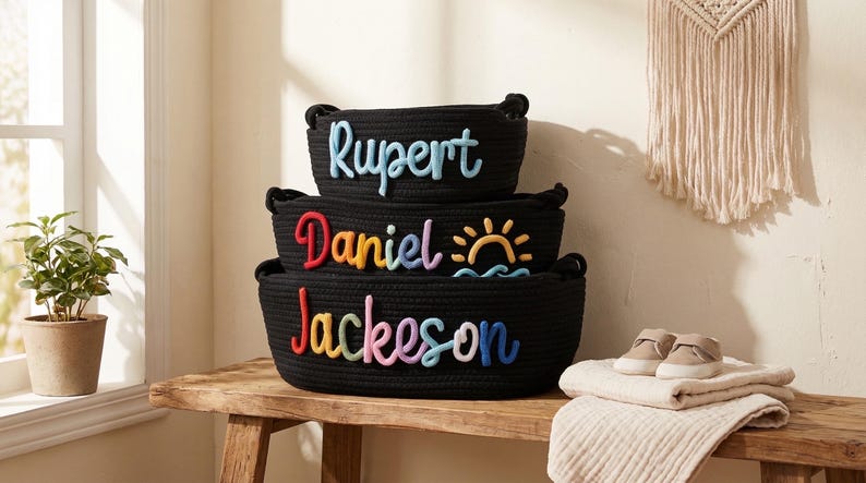 Peut inclure: Trois paniers noirs en corde empil&eacute;s, chacun avec un nom en lettres color&eacute;es : Rupert, Daniel et Jackson. Le panier Daniel a un motif de soleil et de vagues. Une paire de petites chaussures et une couverture pli&eacute;e sont sur la table.
