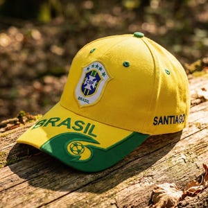 May include: A yellow baseball cap with a green brim and accents, featuring the word "BRASIL" and a shield emblem. The cap also has the word "SANTIAGO" on the side. The cap is resting on a wooden surface.
