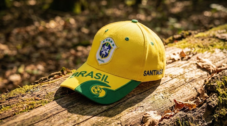 May include: A yellow baseball cap with a green brim and accents, featuring the word "BRASIL" and a shield emblem. The cap also has the word "SANTIAGO" on the side. The cap is resting on a wooden surface.