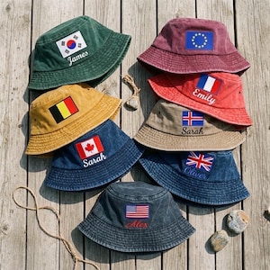 May include: A selection of bucket hats in different colours, each embroidered with a flag and a name. The hats display flags from the USA, UK, Canada, France, South Korea, Belgium, and the European Union. Names are embroidered in a contrasting colour.