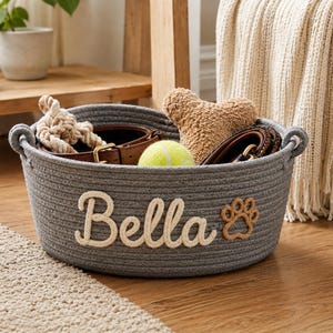 Peut inclure: Un panier gris en corde avec le nom "Bella" en &eacute;criture cr&egrave;me et une empreinte de patte. Il contient un collier pour chien, une balle de tennis, un jouet en corde et un os en peluche. Le panier a des poign&eacute;es.