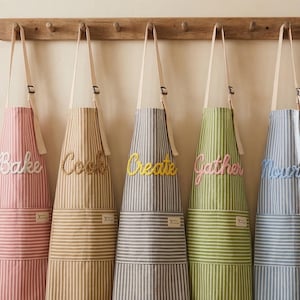 Può includere: Cinque grembiuli a righe appesi a un attaccapanni in legno. Ogni grembiule ha una combinazione di colori diversa e una parola ricamata sul petto: "Bake", "Cook", "Create", "Gather" e "Nourish". I grembiuli hanno una tasca e spalline color crema.