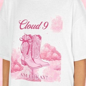 May include: White t-shirt with a pink graphic. The design includes cowboy boots, clouds, and the text "Cloud 9" and "AM I OKAY?". The boots have decorative stitching and bows. The design is set against a pink cloud background.