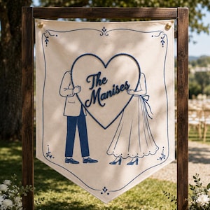 May include: A cream-colored fabric banner suspended from a dark wooden frame, featuring a blue line-art illustration of two figures holding a large heart with the text The Manises embroidered in blue script across the center of the heart.