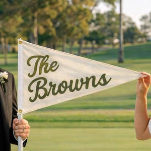 May include: A wedding flag with the text "The Browns" in a script font. The flag is white with green lettering and is held by a person in a black tuxedo and a person in a white wedding dress. The background is a green golf course.