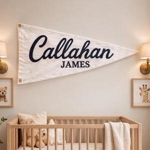May include: A nursery scene with a wooden crib, a personalized pennant banner that reads "Callahan James", and framed animal art. The room is decorated with soft toys, a woven basket, and a fluffy rug, creating a cozy atmosphere.