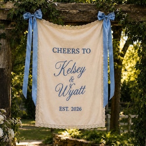 May include: A cream-coloured fabric banner with lace trim and blue ribbon bows, featuring the blue text "CHEERS TO Kelsey & Wyatt EST. 2026" printed in the centre. The banner hangs vertically from a wooden structure in an outdoor setting.