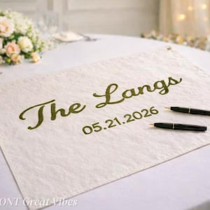 Puede incluir: Alternativa de libro de visitas de lino color crema con las palabras "The Langs" y la fecha "05.21.2026" bordadas en verde. Dos bolígrafos negros están colocados sobre el lino. Un ramo de flores se ve al fondo.