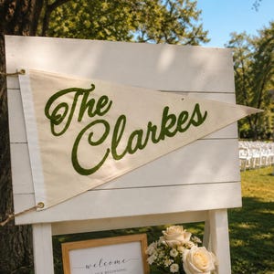 Puede incluir: Un letrero de madera blanco con un banderín que dice "The Clarkes" en letras verdes. Debajo, un letrero enmarcado dice "welcome to our beginning" con las iniciales "g r" y la fecha "06.15.24". Un ramo de rosas y velas completan la exhibición.