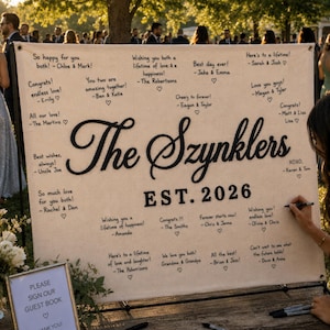 May include: A large, rectangular, off-white fabric guest book sign featuring the black embroidered text The Szynklers and EST. 2026. The fabric is covered in handwritten black ink messages and signatures from wedding guests, each accompanied by a small heart.