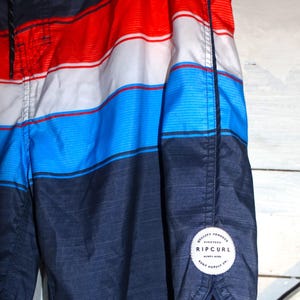 May include: Blue, red, and white striped swim trunks with a black Rip Curl logo patch on the bottom right side. The patch says "Quality Product, Nineteen Sixty Nine, Rip Curl, Surf Supply Co."