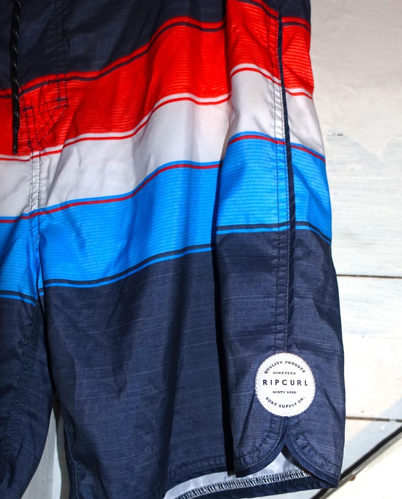 May include: Blue, red, and white striped swim trunks with a black Rip Curl logo patch on the bottom right side. The patch says "Quality Product, Nineteen Sixty Nine, Rip Curl, Surf Supply Co."