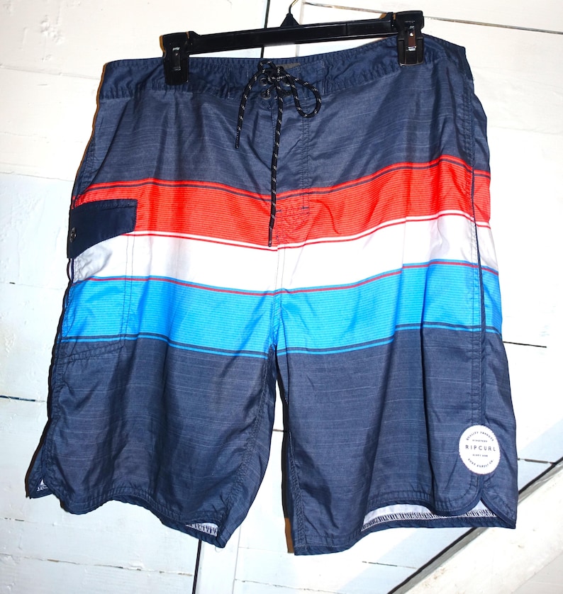 May include: Blue, red, and white striped swim trunks with a black drawstring and a Rip Curl logo on the side.
