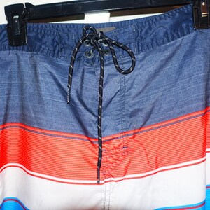 May include: Blue, red, and white striped swim trunks with a black drawstring. The trunks have a small pocket on the left side.