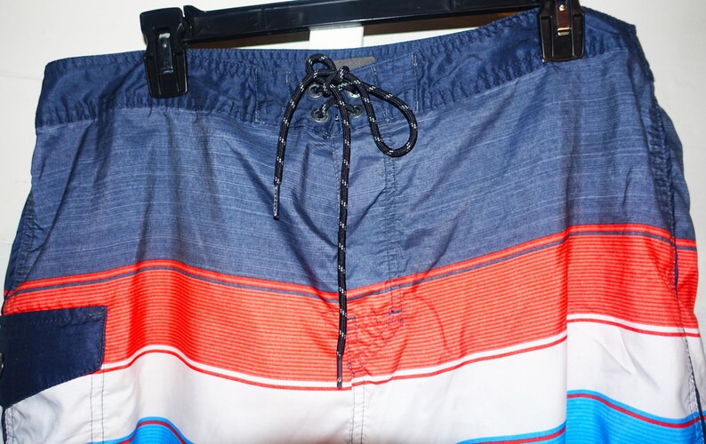 May include: Blue, red, and white striped swim trunks with a black drawstring. The trunks have a small pocket on the left side.