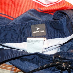 May include: Blue and red striped swim trunks with a Rip Curl logo tag. The tag is black with white text and a white wave logo. The fabric is 100% polyester.