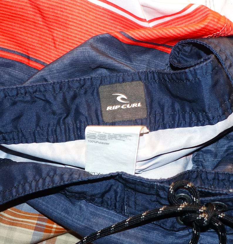 May include: Blue and red striped swim trunks with a Rip Curl logo tag. The tag is black with white text and a white wave logo. The fabric is 100% polyester.