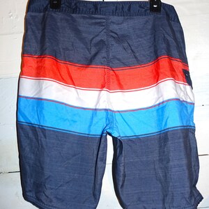 May include: Blue, red, and white striped swim trunks with a dark blue background. The trunks have a drawstring waist and a back pocket.