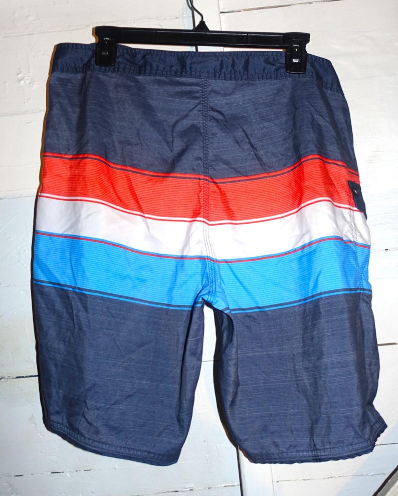 May include: Blue, red, and white striped swim trunks with a dark blue background. The trunks have a drawstring waist and a back pocket.