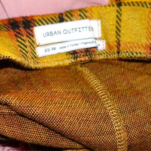 Puede incluir: Tela de cuadros amarilla y naranja con una etiqueta blanca que dice "URBAN OUTFITTER" y "XS-TP".