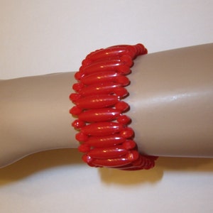 May include: A vibrant red bracelet composed of numerous, glossy, elongated beads. The beads are strung together, forming a flexible band that wraps around the wrist. The bracelet's design is simple yet eye-catching, with a bold, solid color.