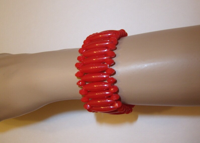 May include: A vibrant red bracelet composed of numerous, glossy, elongated beads. The beads are strung together, forming a flexible band that wraps around the wrist. The bracelet's design is simple yet eye-catching, with a bold, solid color.