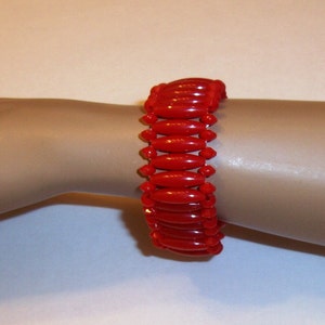 May include: A vibrant red stretch bracelet. The bracelet is composed of numerous elongated, oval-shaped beads. The beads are strung together, creating a flexible and eye-catching accessory. The bracelet is shown on a mannequin arm.