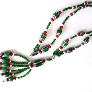 May include: A beaded necklace featuring green, red, and white beads. The necklace has a tassel of green beads and a clasp. The design includes elongated green and white striped beads, and small red and white beads.