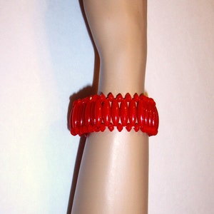 May include: A vibrant red bracelet with an oval bead design. The bracelet is made of glossy, polished beads strung together. The jewelry is worn on a mannequin arm, showcasing the bracelet's color and style. A fashion accessory.