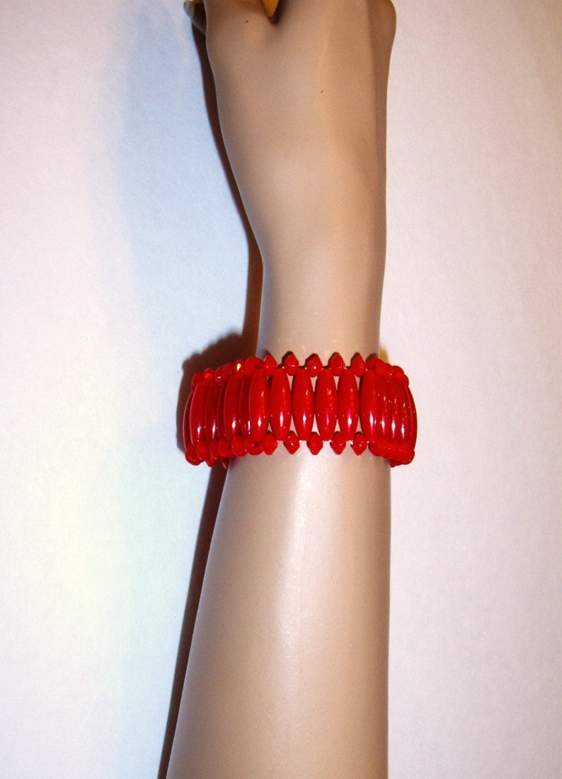 May include: A vibrant red bracelet with an oval bead design. The bracelet is made of glossy, polished beads strung together. The jewelry is worn on a mannequin arm, showcasing the bracelet's color and style. A fashion accessory.